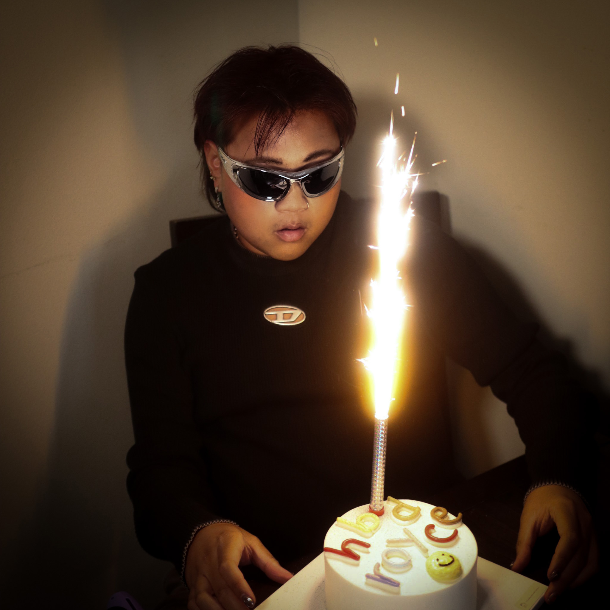 Person in silver Gentle Monster sunglasses lit by a sparkler candle in a dark room, the edited hero frame for the Elsewhere campaign.